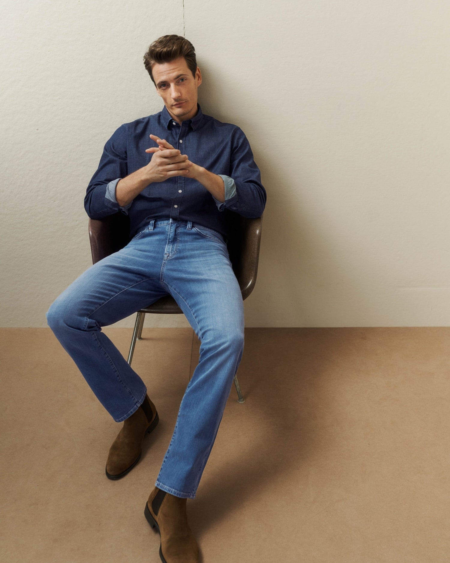 A man sits in a chair against a plain wall, wearing a dark blue button-up shirt, light blue jeans, and brown boots.