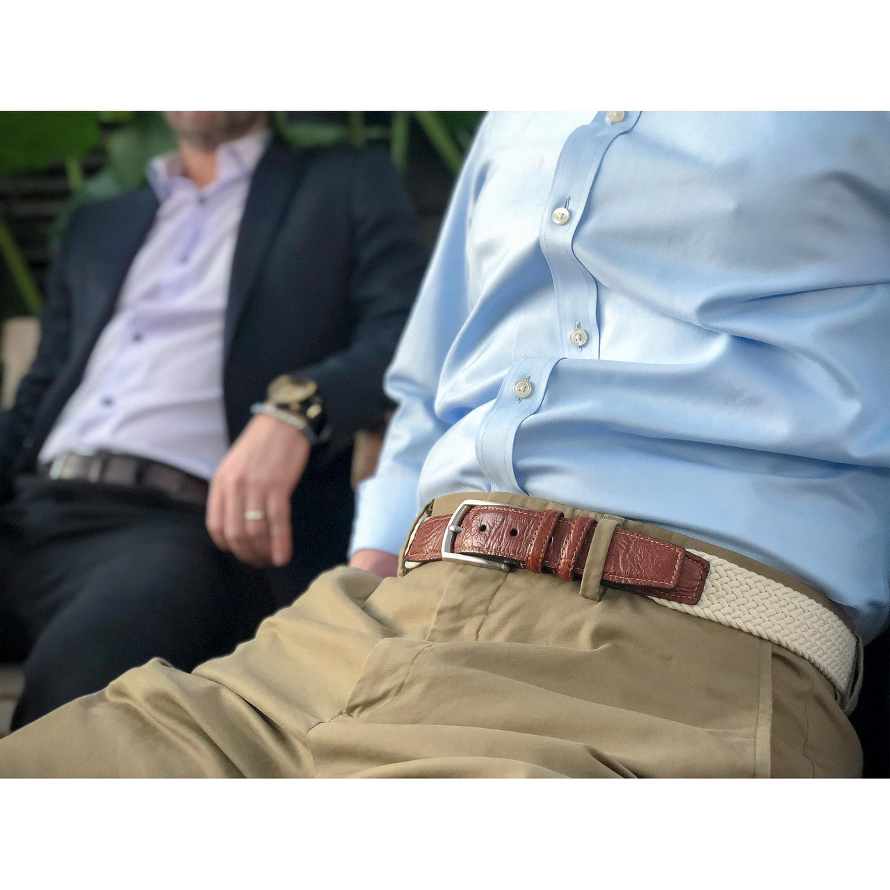 Two men seated; one in a light blue dress shirt and khaki pants with a brown belt in the foreground, another in a dark suit and lavender shirt in the background.