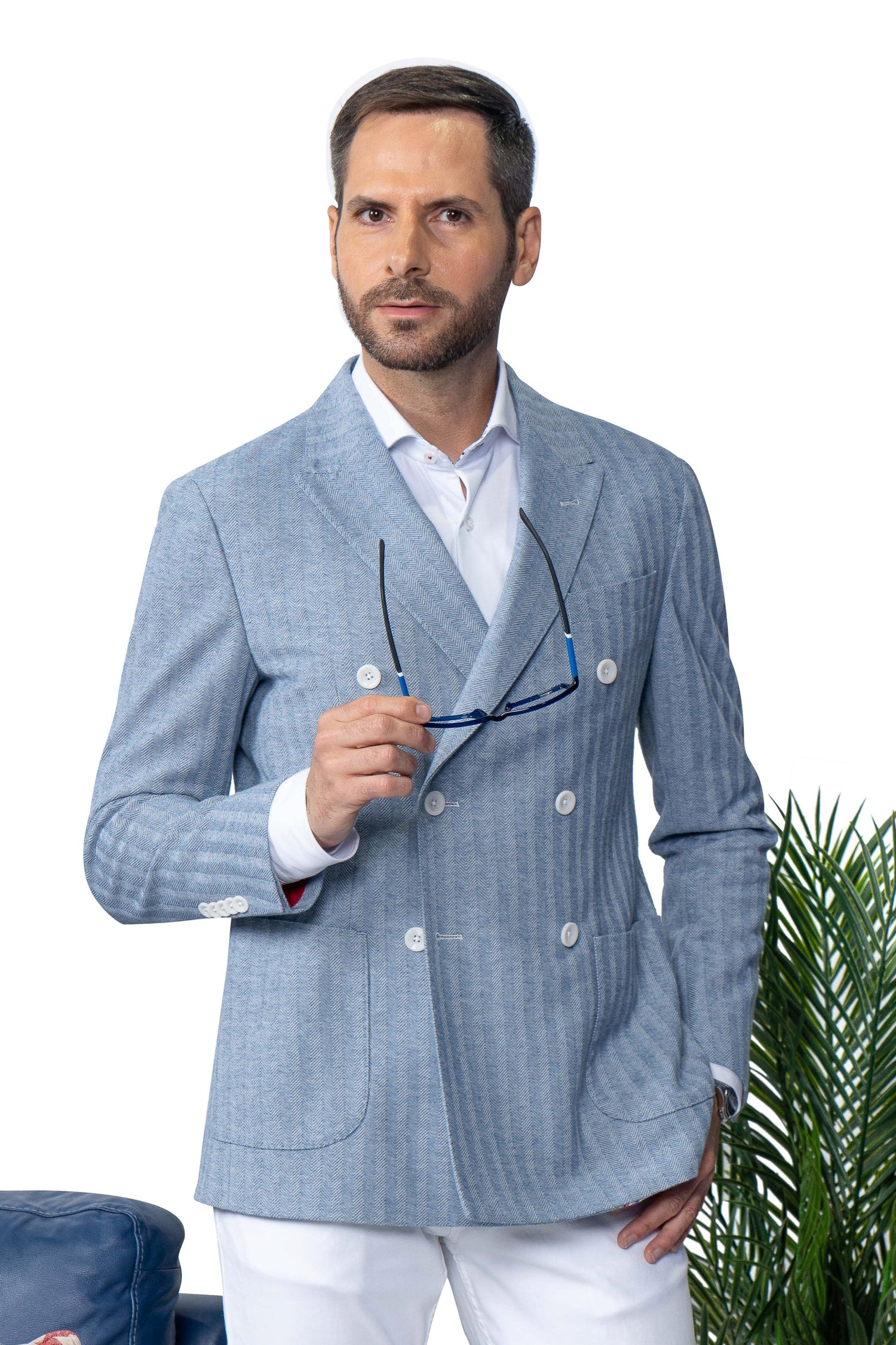 A man in a light blue double-breasted blazer and white pants holds eyeglasses in one hand, standing next to a green plant against a white background.