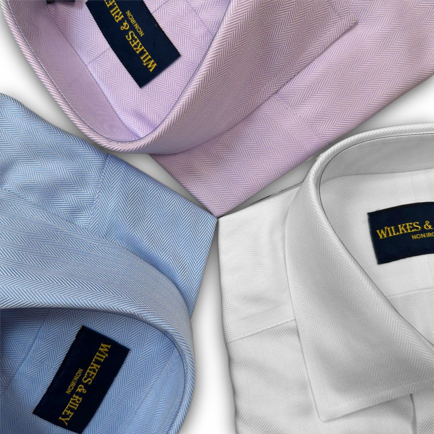 Three herringbone-pattern dress shirts in light pink, light blue, and white, each with a Wilkes & Riley label on the inside collar, arranged on a flat surface.