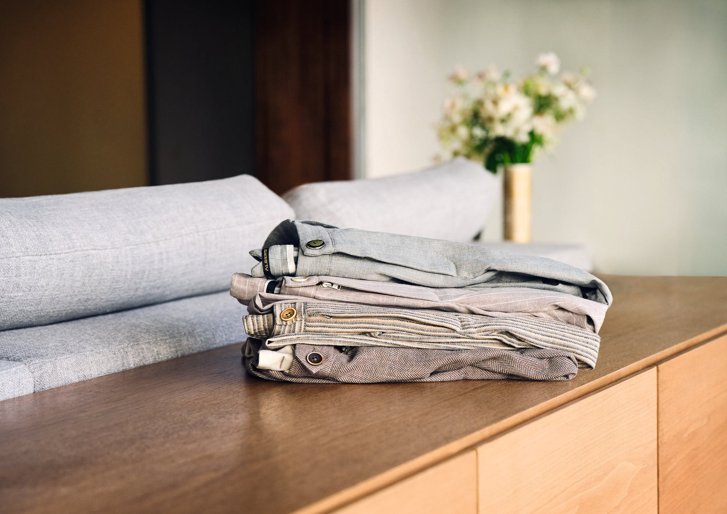 A neatly folded stack of light-colored pants sits on a wooden surface in front of a gray couch, with a vase of flowers in the background.