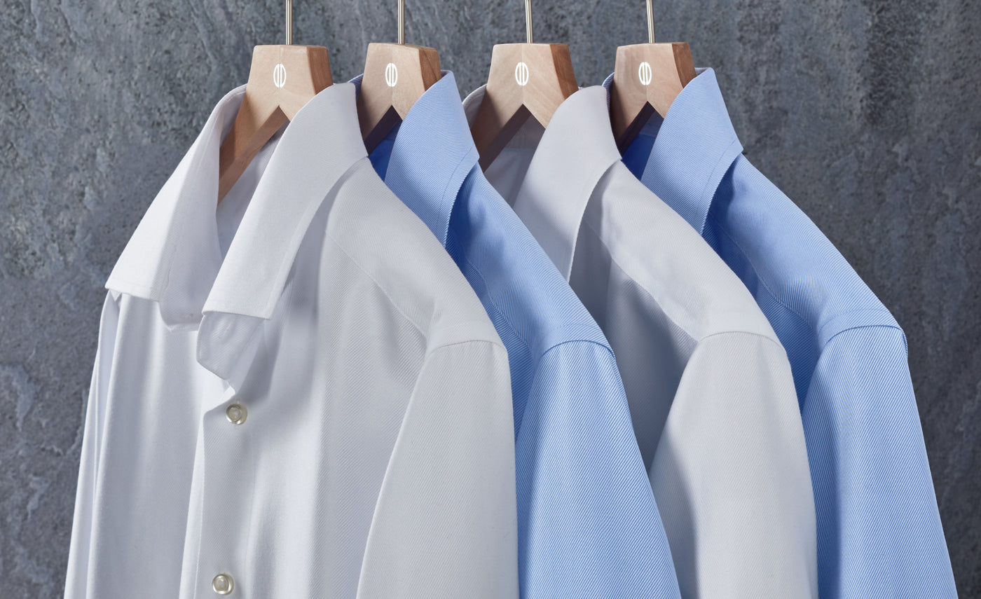 Four collared dress shirts, two white and two light blue, hang neatly on wooden hangers against a gray textured background.