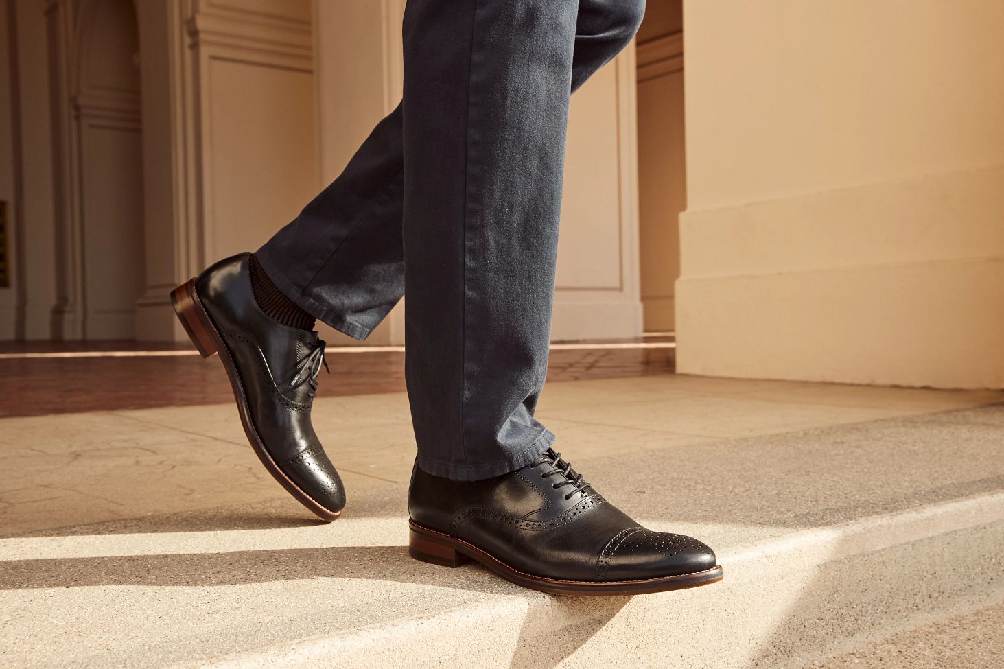 A person wearing dark dress shoes and navy pants steps up onto a light-colored stone surface in an outdoor setting.