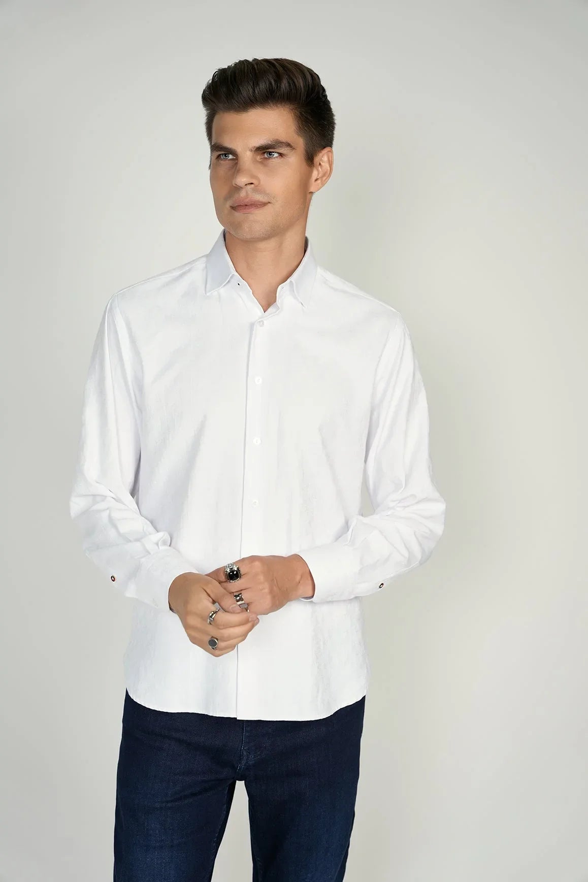 A man stands against a plain background wearing a white button-up shirt and dark blue jeans, with his hands loosely clasped.