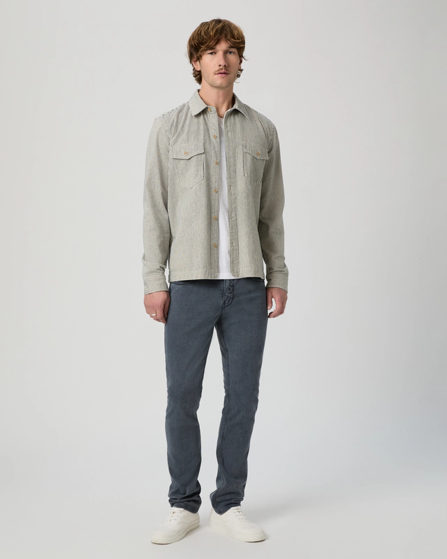 A man stands facing forward, wearing a light gray button-up shirt over a white t-shirt, dark gray jeans, and white sneakers, against a plain light background.