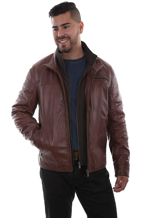 Man standing and smiling, wearing a brown leather jacket over a navy blue shirt and black pants against a plain white background.