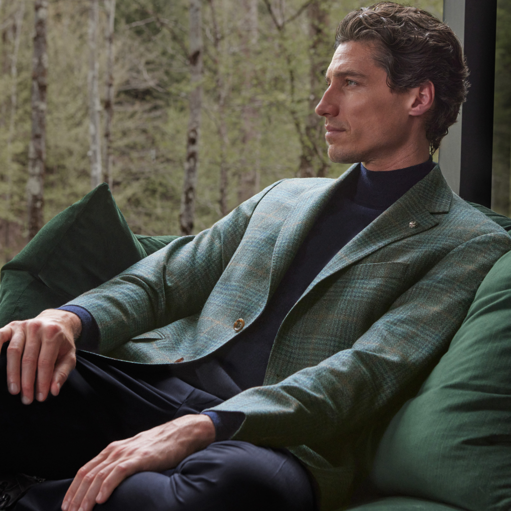 A man in a green plaid blazer and black turtleneck sits on a green couch, looking to the side, with a forest visible through the window behind him.