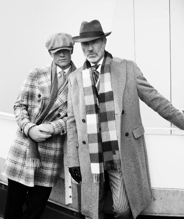 Two men wearing long coats, scarves, and hats stand side by side; one leans slightly while the other stands upright. The photo is in black and white.