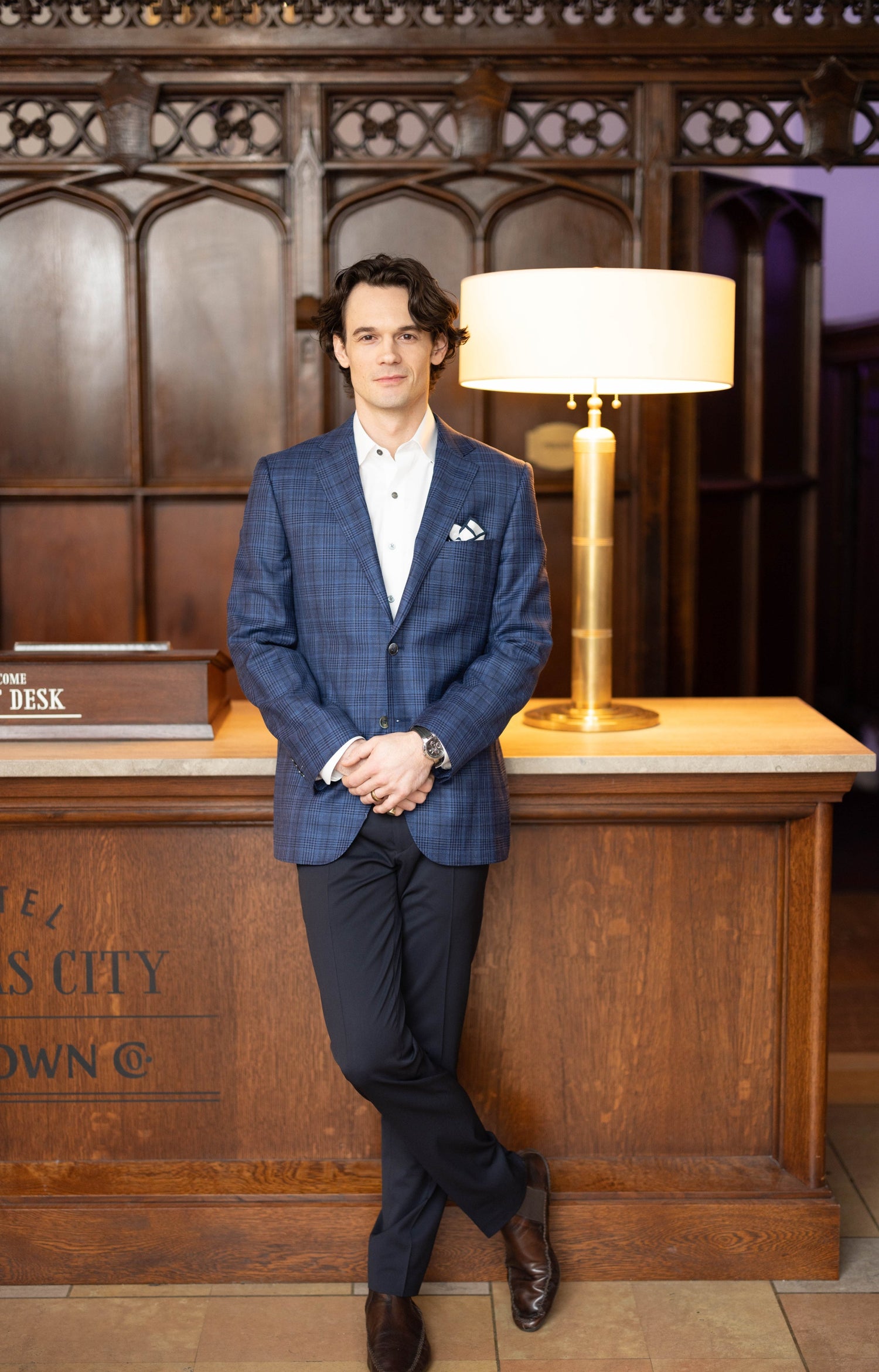 A man in a blue blazer and dark pants stands in front of a wooden reception desk with a lamp, posing with one leg crossed over the other.