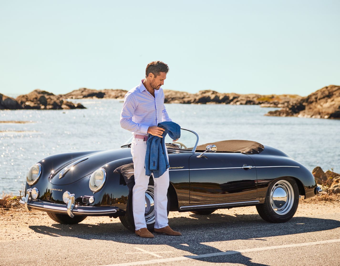 A man stands beside a classic black convertible parked near water, holding a blue jacket, with rocky shoreline and clear sky in the background.