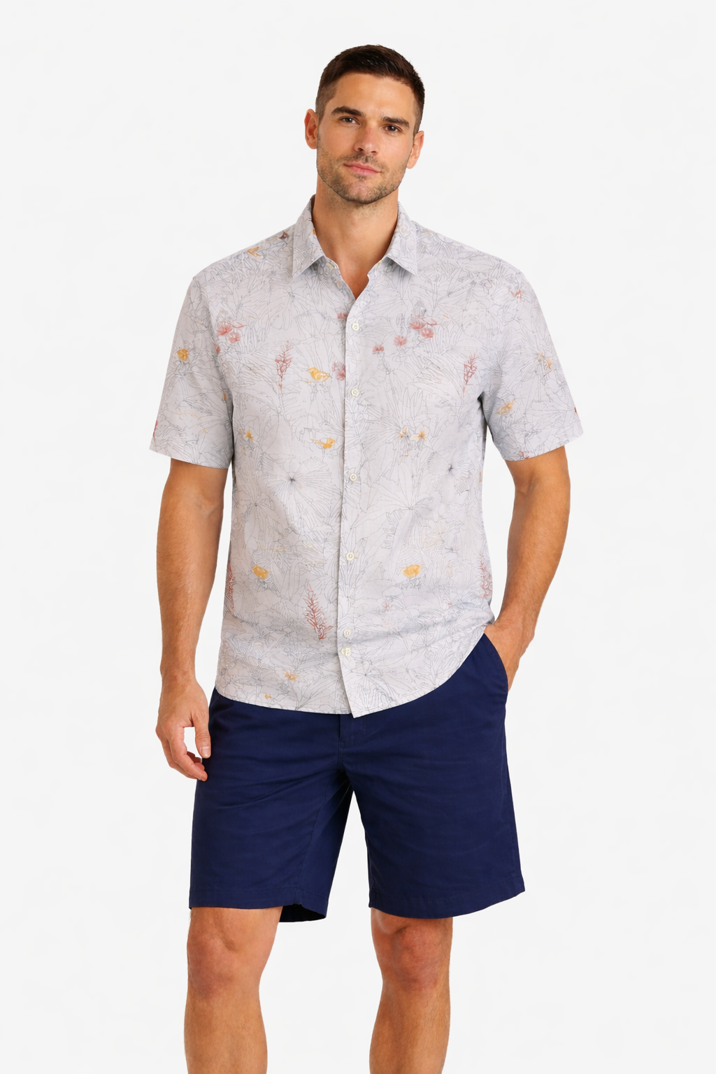 A man stands wearing a light-colored, short-sleeve, button-up shirt with a subtle floral pattern and dark blue shorts against a plain white background.