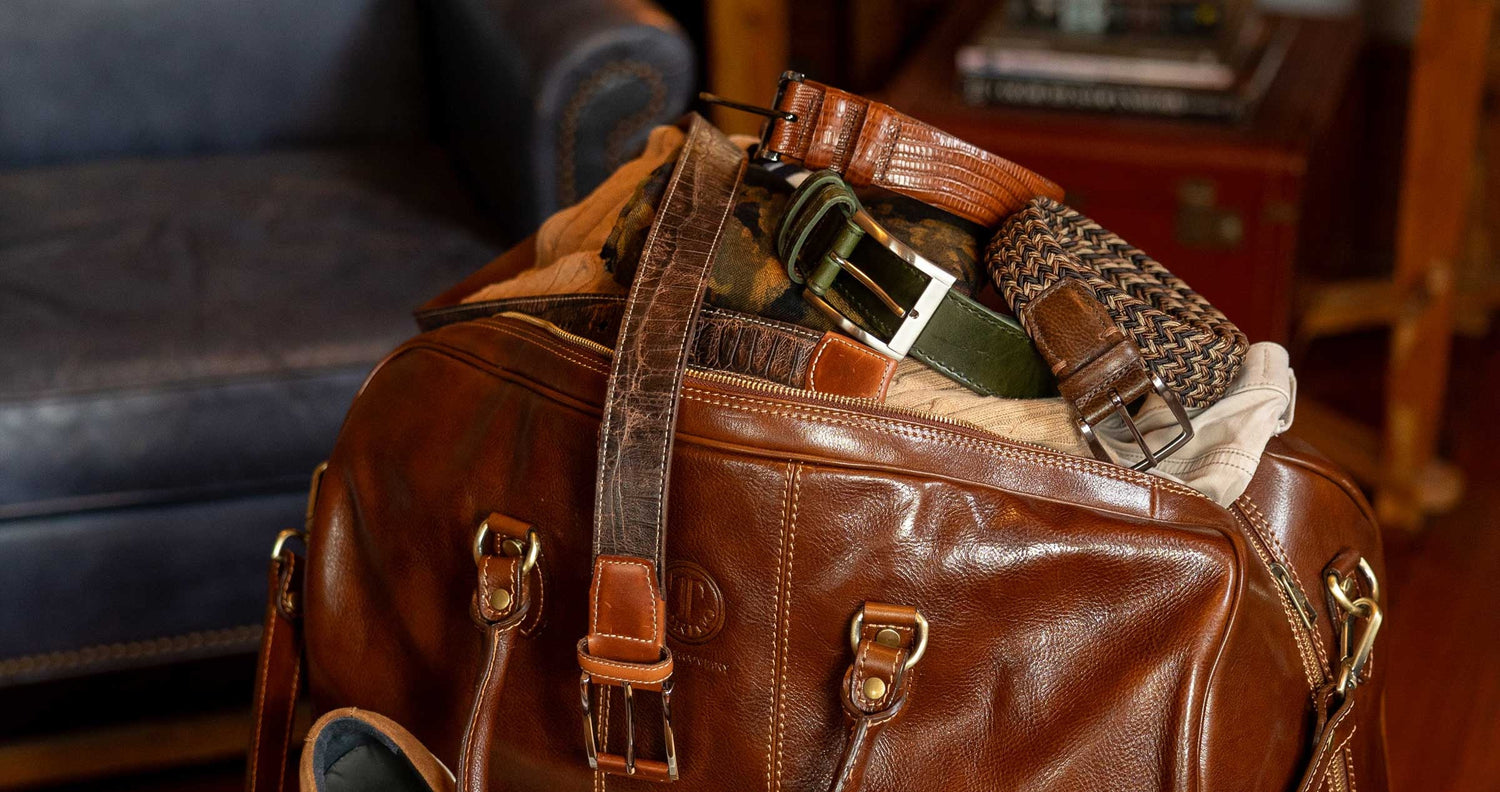 A brown leather duffel bag filled with belts, clothing, and accessories sits next to a blue armchair in a warmly lit room.