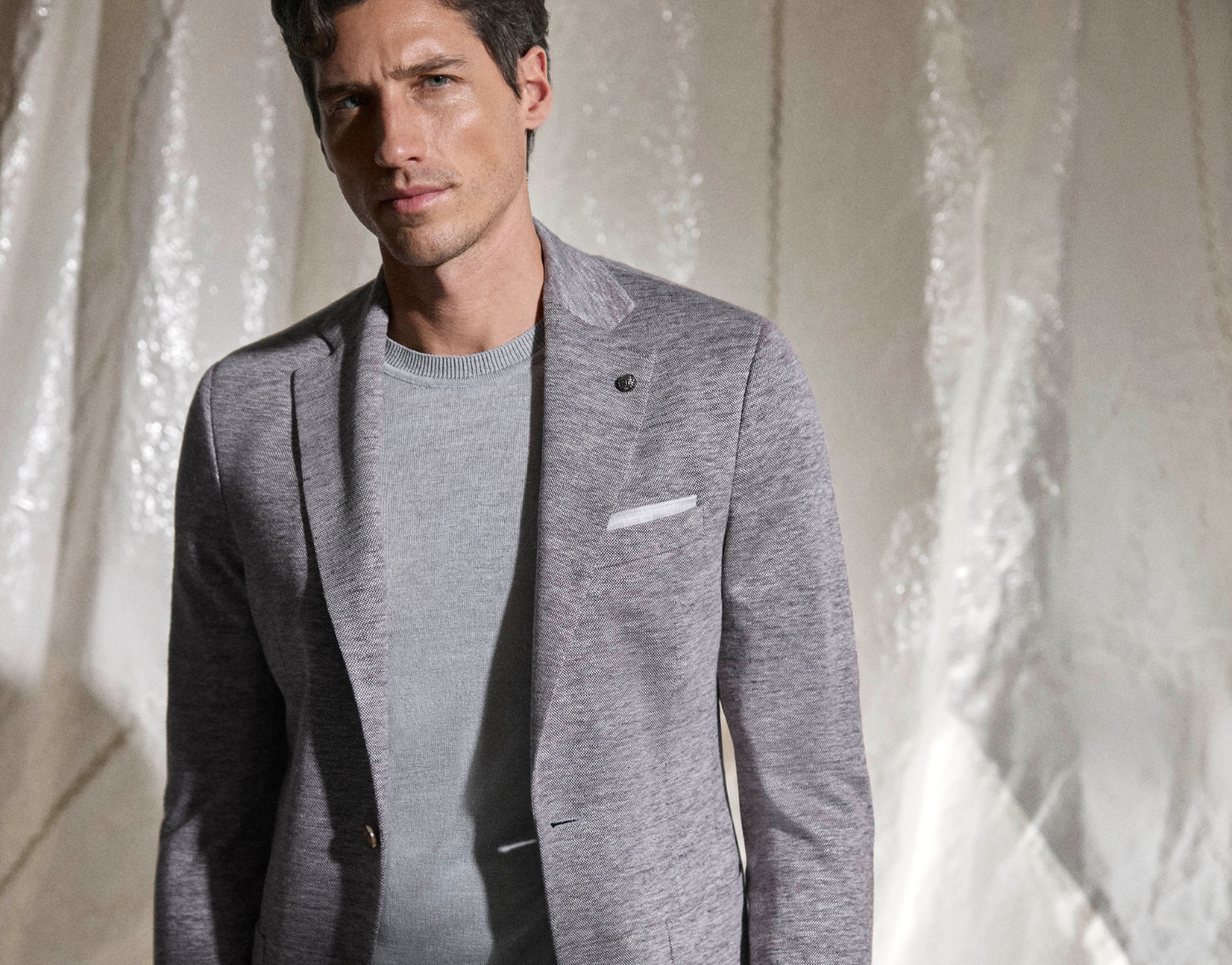 A man wearing a light gray blazer over a gray sweater stands in front of a light-colored, textured backdrop.