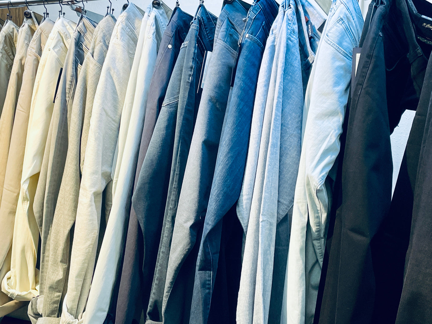 A row of pants in various colors and denim washes hanging on metal hangers on a clothing rack.