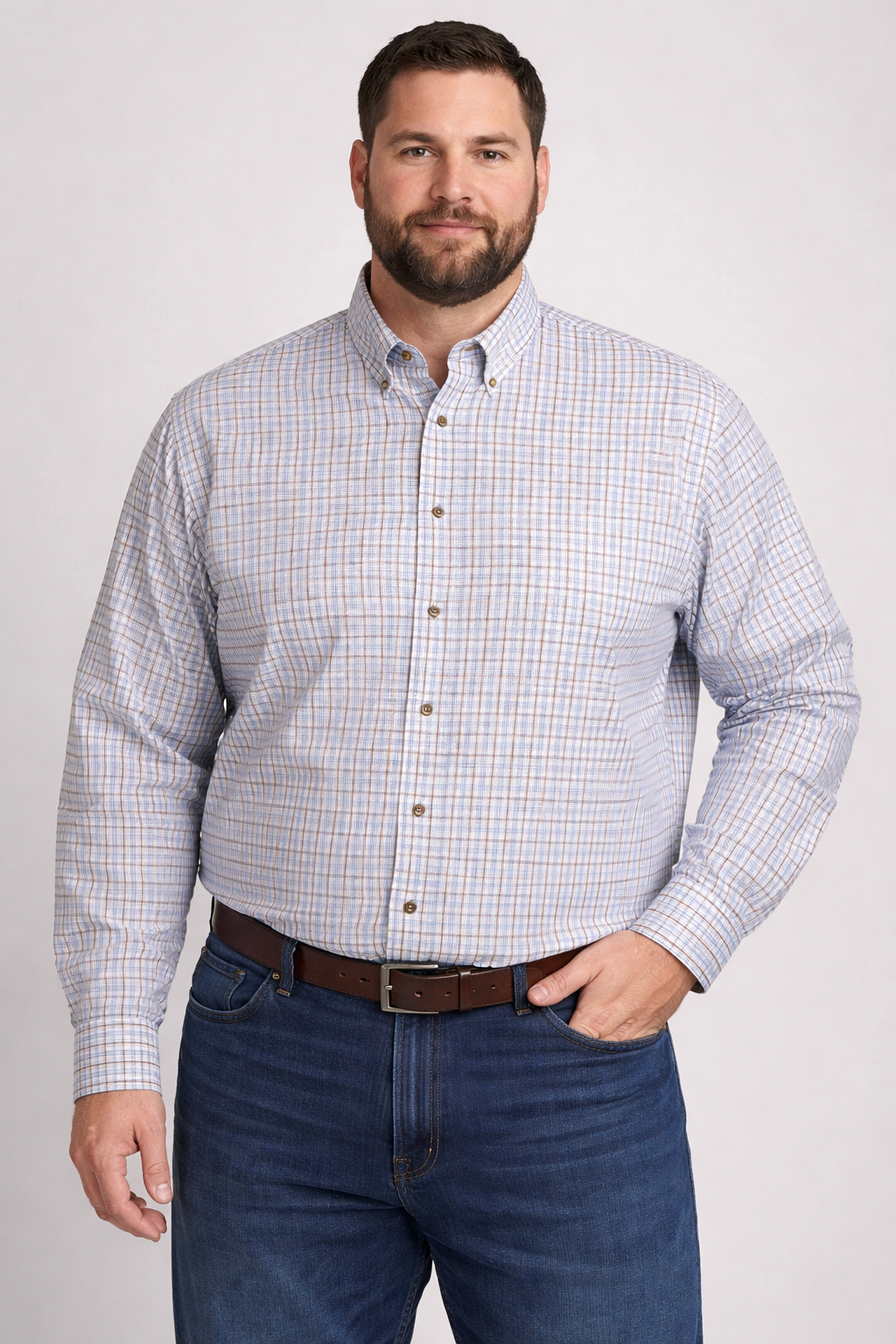 Man standing and smiling, wearing a light blue and tan plaid button-up shirt with blue jeans and a brown belt against a neutral background.