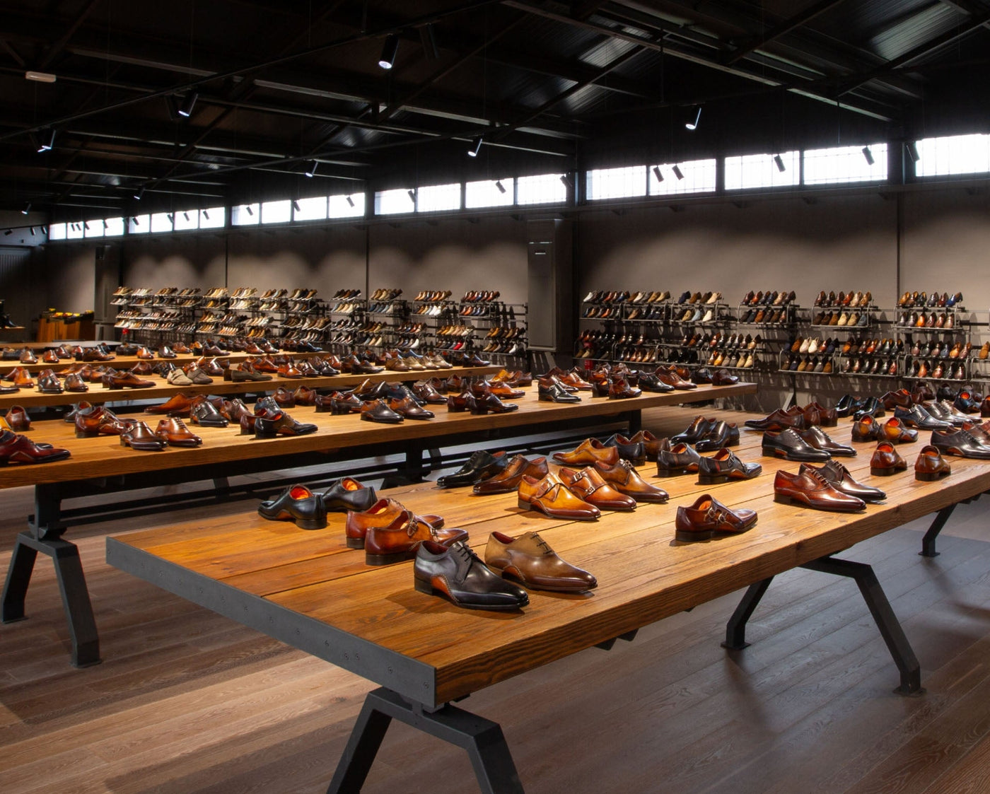 A spacious shoe store interior displays rows of polished mens dress shoes on wooden tables and shelves under bright lighting.