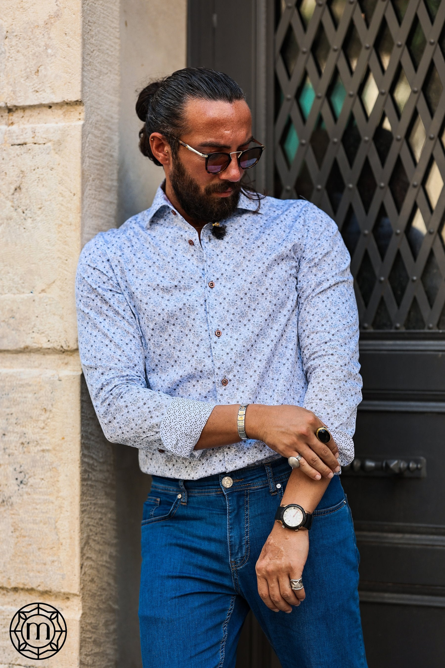 A man with a beard and sunglasses stands against a wall, rolling up the sleeve of his patterned shirt, wearing blue jeans, a ring, a watch, and a bracelet.