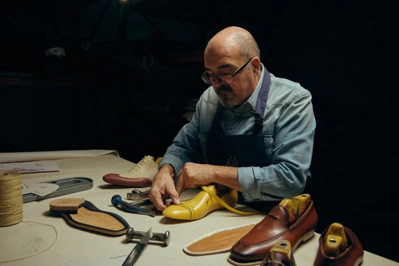 A man wearing glasses and an apron works on crafting a yellow leather shoe at a workbench, with tools and completed shoes around him.