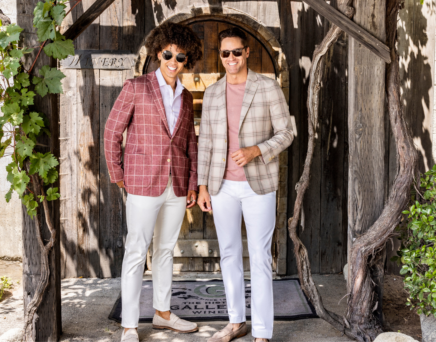 Two men wearing sunglasses, plaid blazers, and white pants stand and smile in front of a rustic wooden doorway with grapevines on either side.