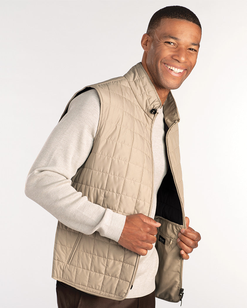 A man smiles while wearing a light beige quilted vest over a light gray long-sleeve shirt. He is holding the open front of the vest with one hand.