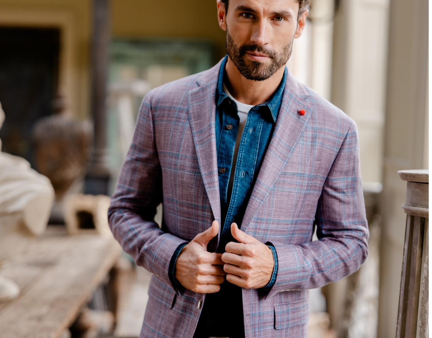A man wearing a light purple plaid suit jacket over a blue denim shirt stands indoors, holding the edges of his jacket with both hands.