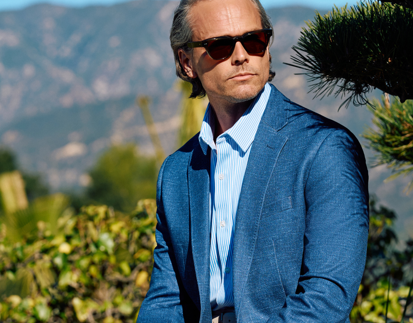 A man wearing sunglasses and a blue suit jacket over a striped shirt sits outdoors with greenery and mountains in the background.