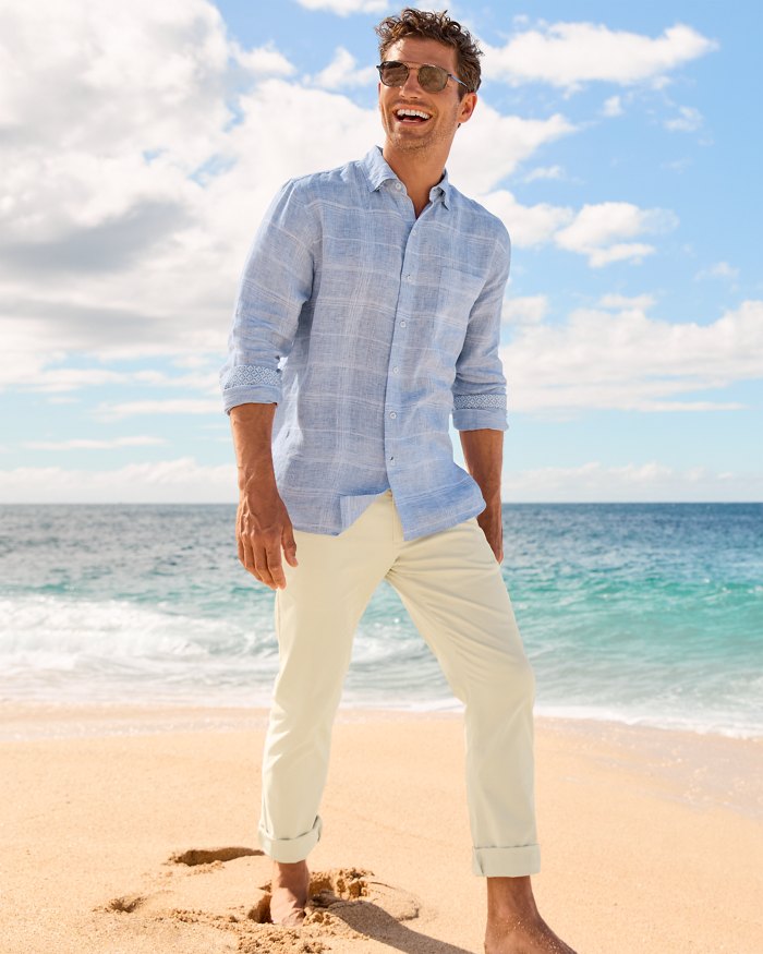 A man wearing sunglasses, a light blue button-down shirt, and rolled-up light-colored pants stands barefoot on a sandy beach with the ocean and blue sky in the background.