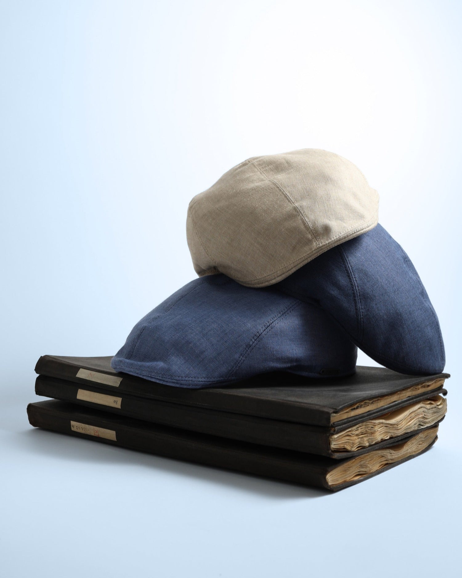 Two flat caps, one beige and one blue, are stacked on three old, hardcover books with visible worn pages, set against a plain light background.