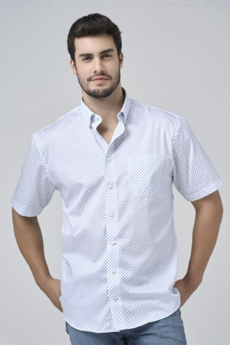 A man with short dark hair and a beard wears a white, short-sleeved, button-up shirt with a subtle dot pattern and blue jeans, standing against a plain light background.