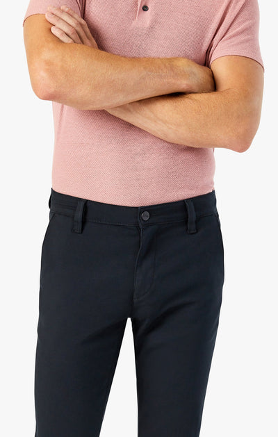 A person wearing slim 34 Heritage Black High-Flyer trousers and a short-sleeve pink polo shirt stands with arms crossed, shown from shoulders to mid-thigh against a plain background.