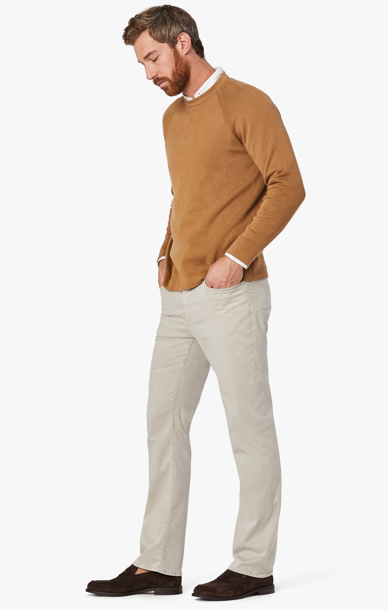 A bearded man stands with hands in pockets, wearing a tan sweater over a white shirt, 34 Heritage Dawn Twill light beige straight-leg pants, and dark brown shoes against a plain white background.