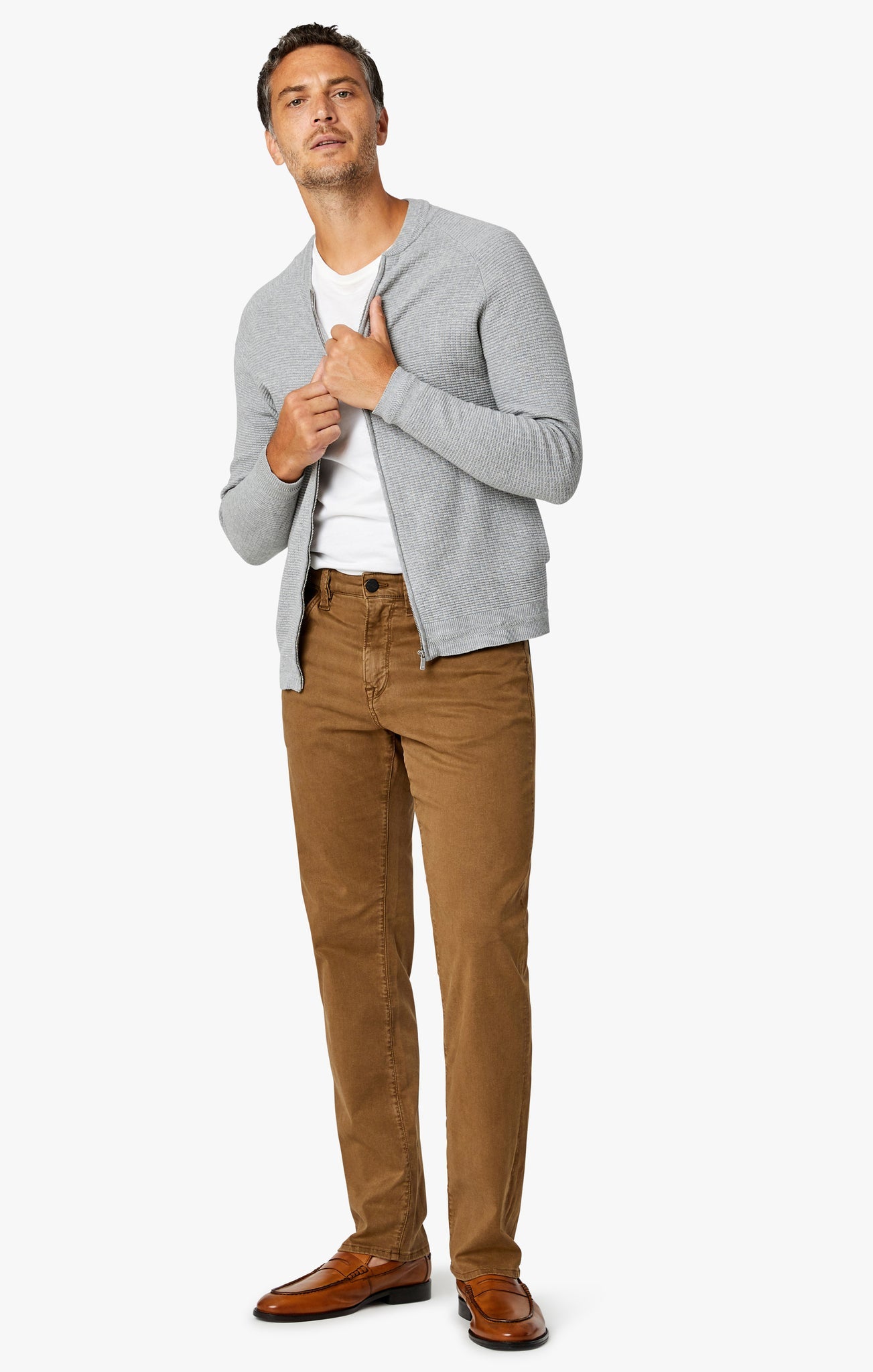 A man faces forward against a plain white background, wearing a light gray zip-up sweater, white t-shirt, 34 Heritage Tobacco Twill straight-leg pants with a comfort rise, and brown dress shoes.