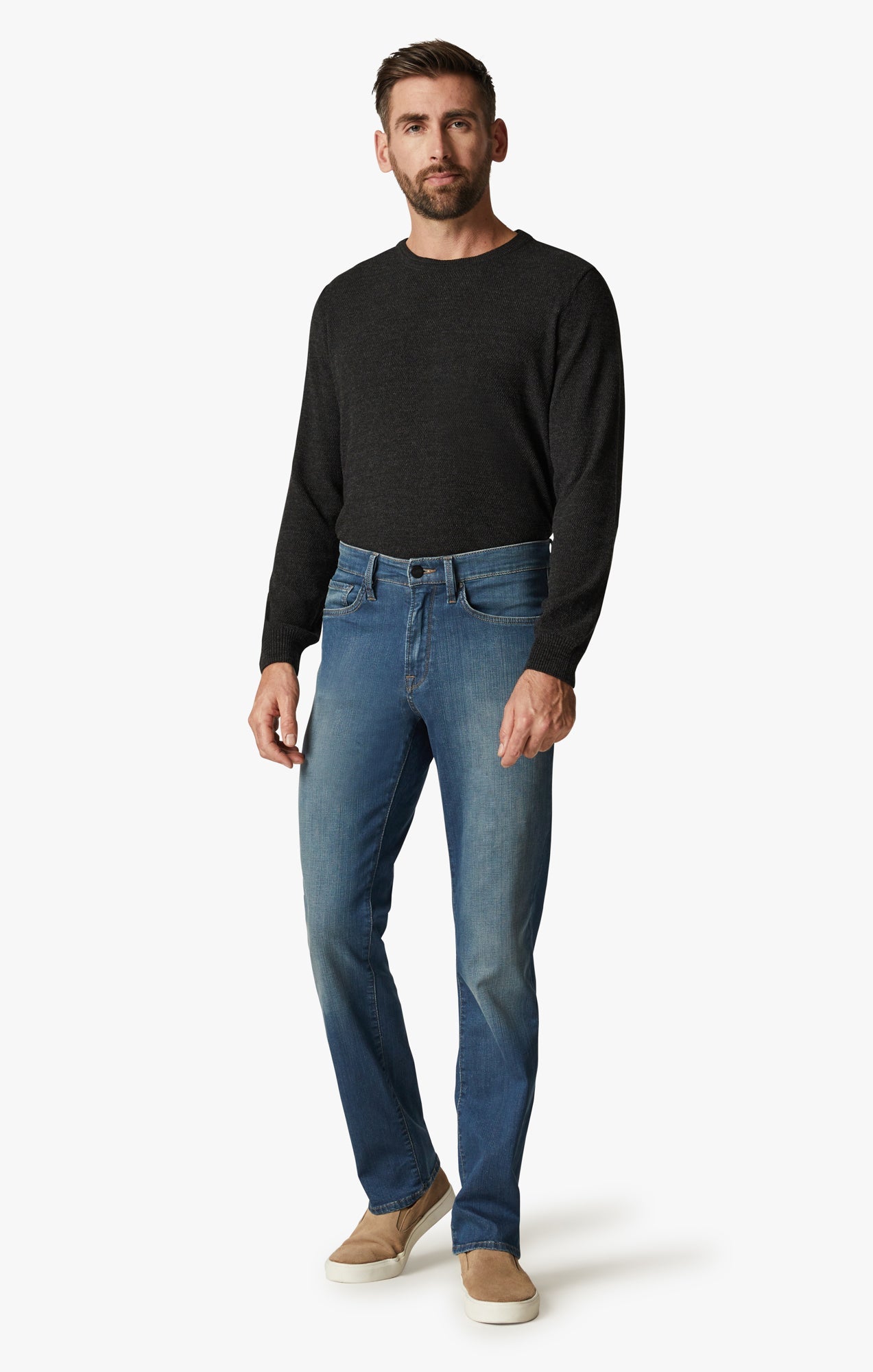 A man stands in a relaxed pose wearing 34 Heritage Mid Cashmere soft denim straight leg jeans in light blue, paired with a black long-sleeve sweater and beige slip-on shoes against a plain white background.
