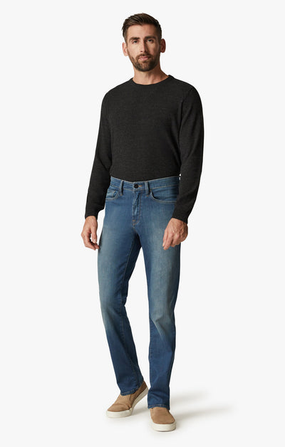 A man stands in a relaxed pose wearing 34 Heritage Mid Cashmere soft denim straight leg jeans in light blue, paired with a black long-sleeve sweater and beige slip-on shoes against a plain white background.
