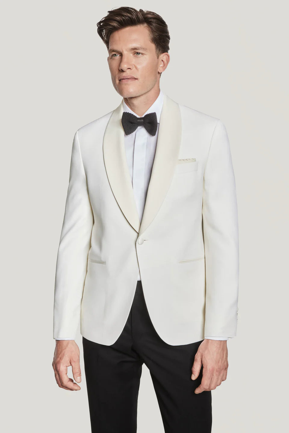 A man wearing the Jack Victor Ethan Cream Dinner Jacket by Jack Victor, paired with a white dress shirt, black bow tie, and black pants, stands against a plain background.