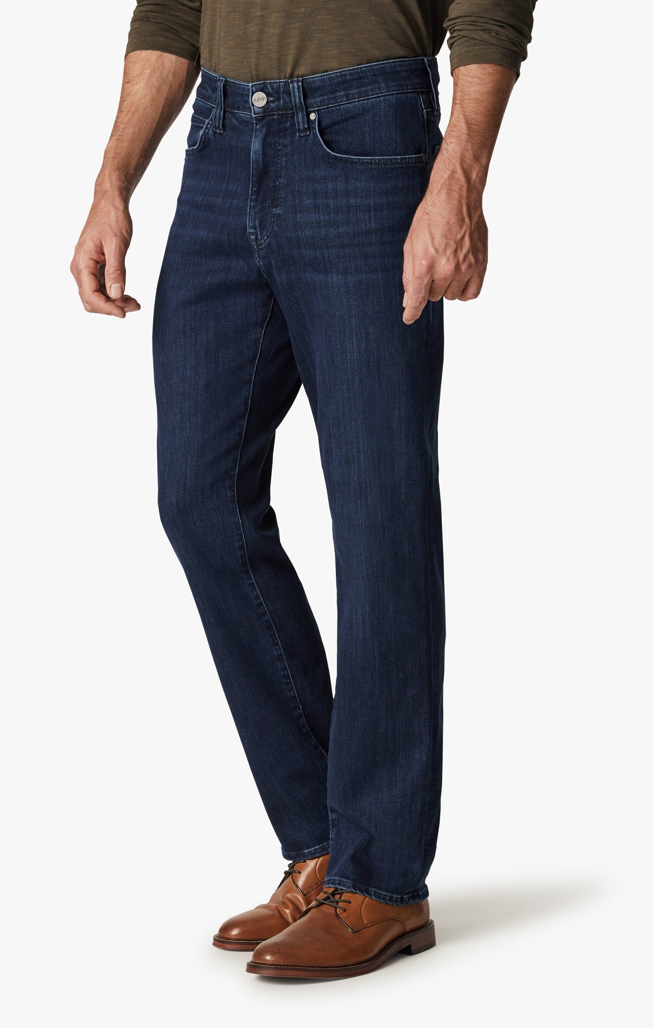 A man wearing 34 Heritage Dark Midnight Brushed Urban jeans from 34 Heritage, a long-sleeve olive shirt, and brown leather dress shoes stands against a plain background.