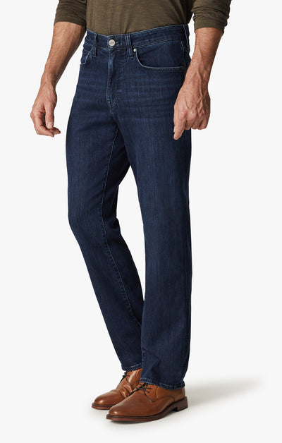 A man wearing 34 Heritage Dark Midnight Brushed Urban jeans from 34 Heritage, a long-sleeve olive shirt, and brown leather dress shoes stands against a plain background.