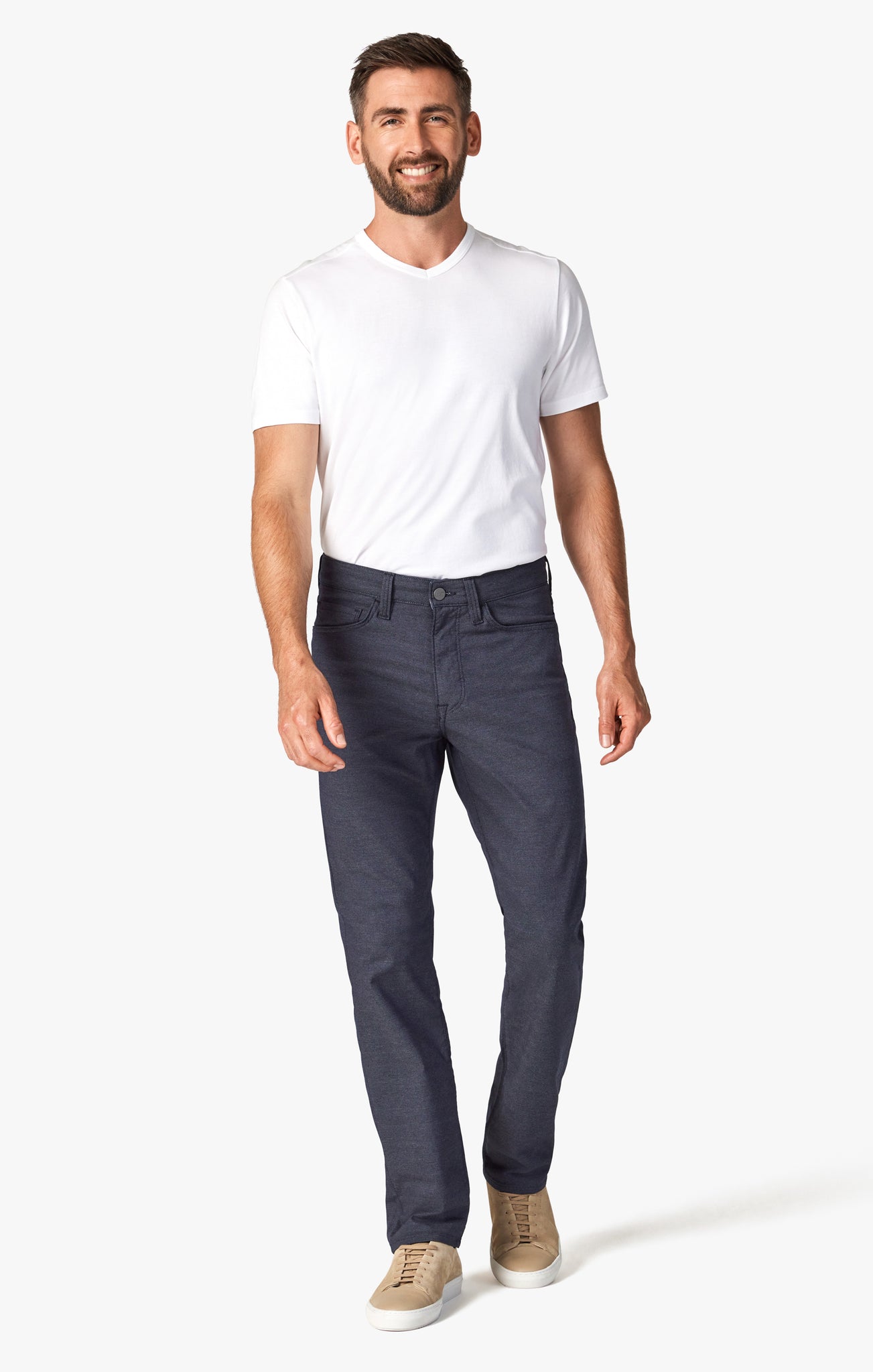 A man stands and smiles against a plain white background, wearing a white t-shirt, beige sneakers, and 34 Heritage Navy Coolmax denim pants by 34 Heritage.