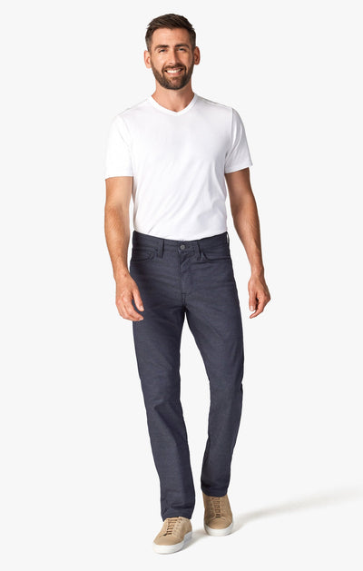 A man stands and smiles against a plain white background, wearing a white t-shirt, beige sneakers, and 34 Heritage Navy Coolmax denim pants by 34 Heritage.