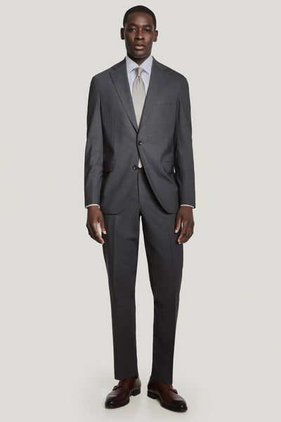 A man stands facing forward in the Jack Victor Solid Wool Suit by Jack Victor, paired with a light blue shirt, light tie, and brown dress shoes. The suit’s wrinkle-resistant fabric ensures a polished look against a plain light background.