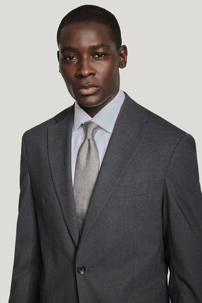 A man stands against a plain light background wearing the Jack Victor Solid Wool Suit in dark grey, paired with a light grey tie and white dress shirt by Jack Victor.