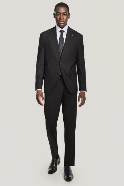 A man stands facing forward, wearing a Jack Victor Solid Wool Suit with a white dress shirt, black tie, and black dress shoes against a plain light background.