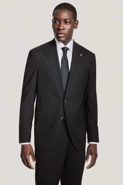 A man in a Jack Victor Solid Wool Suit by Jack Victor, paired with a white dress shirt and black tie, stands against a plain light background looking slightly to the side.