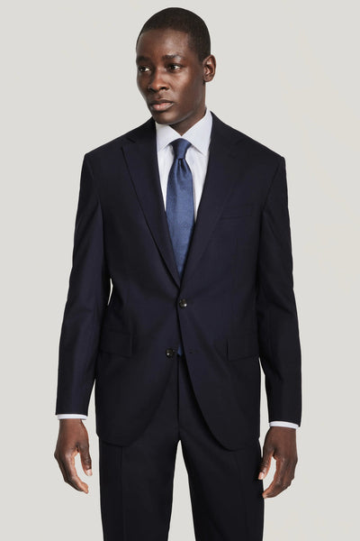 A man in a Jack Victor Solid Wool Suit, featuring wrinkle-resistant stretch wool in navy, paired with a white dress shirt and blue tie, stands against a plain light background.