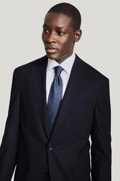 A man wears the Jack Victor Solid Wool Suit by Jack Victor, featuring navy wrinkle-resistant stretch wool, paired with a white dress shirt and blue tie, standing against a plain light background.