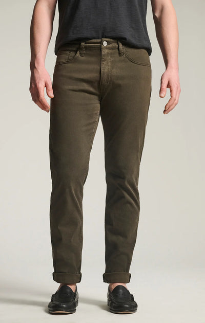 A person stands facing forward against a plain background, wearing 34 Heritage Courage Twill Pants in olive green, paired with a dark t-shirt and black shoes.