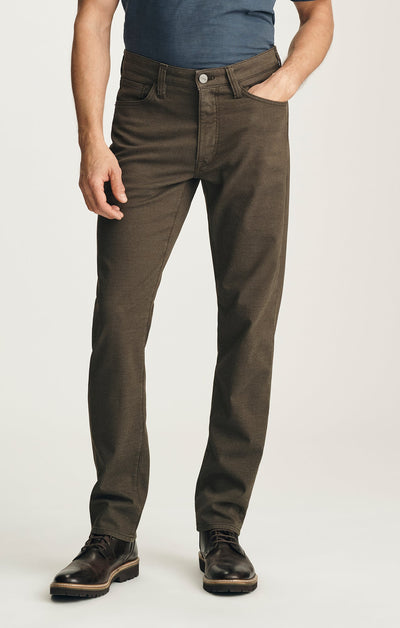 A man wearing a blue 34 Heritage Courage Coolmax t-shirt and straight-leg, dark olive-green dress pants with brown leather boots.