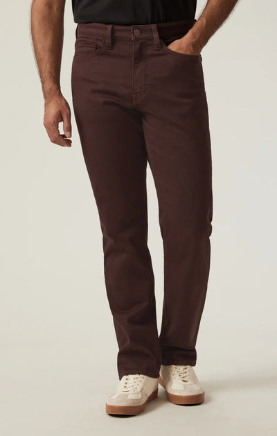 Wearing 34 Heritage Charisma Diagonal Big&Tall burgundy twill pants, a black shirt, and white sneakers, a person stands with one hand in their pocket against a plain background.