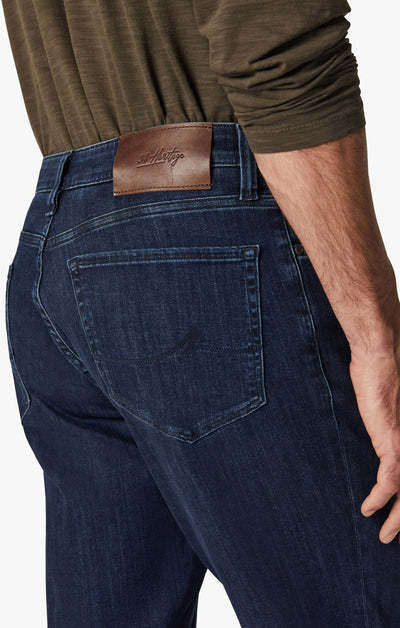 Seen from the back, a person wears 34 Heritage Dark Midnight Brushed Urban straight-leg jeans with a brown leather waistband patch and an olive green long-sleeve shirt.