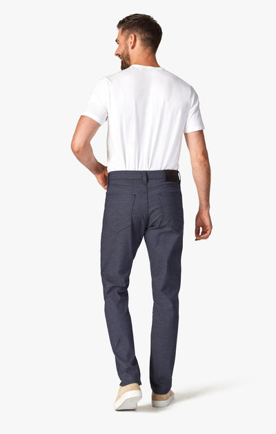 A man stands with his back to the camera, wearing a white t-shirt and 34 Heritage Navy Coolmax jeans by 34 Heritage, paired with light-colored shoes against a plain white background.